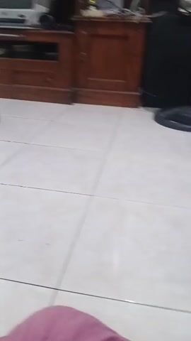 The video shows a person sitting on the floor and holding a small plastic bag. The bag appears to contain some sort of medical supplies, possibly medication or bandages, with labels in a foreign language. The person is wearing traditional clothing. In the background, there is a bed and furniture. Another person is partially visible in the background, wearing a red shirt and black shorts, and there is a fan. The setting appears to be a residential interior.