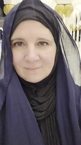 A selfie video shows a woman wearing a navy blue hijab and a black under-scarf, smiling at the camera. Behind her, in the background, is a grand hall with tall, ornate white columns topped with gold. Several people, including children, are walking away from the camera down the hall. The hall appears to be a place of worship or significant cultural importance due to its elaborate architecture.