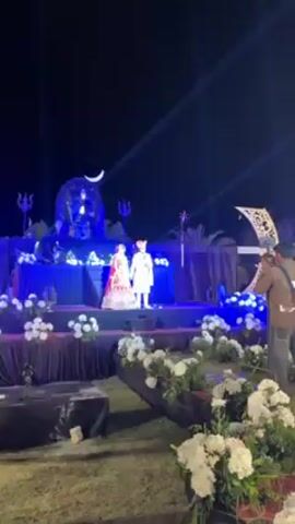 The video shows a couple standing on a stage, likely at a wedding reception. The bride is wearing a red and gold lehenga, and the groom is in a white sherwani. They are both wearing garlands. Behind them is a large statue lit in blue light, featuring a crescent moon above it. There are white floral arrangements on the stage and in front of it, with a photographer standing on the right side of the scene. The background is dark, with spotlights shining upwards. The scene is outdoors. The couple is looking at each other.