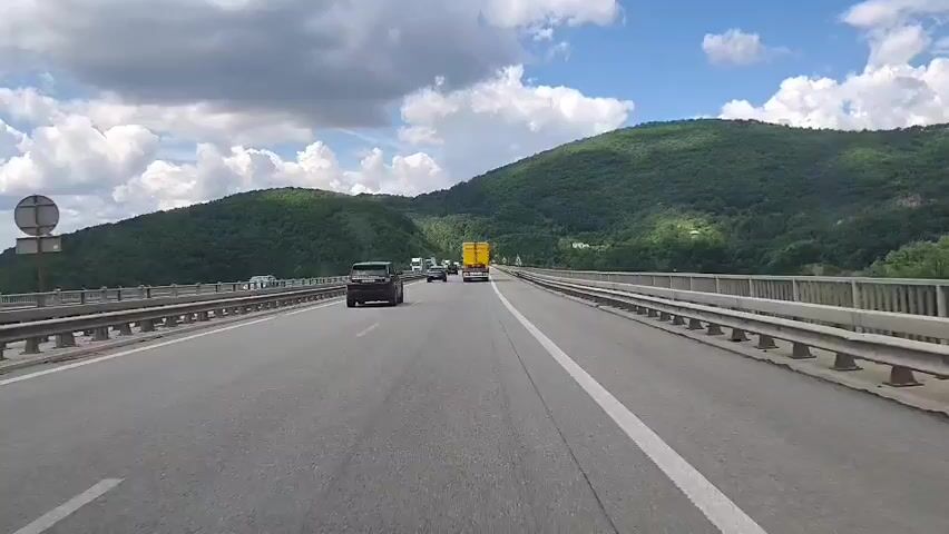 This video shows a first-person perspective of driving on a highway bridge, likely in Vrachesh, Bulgaria, given the provided context. The highway has multiple lanes and is lined with metal guardrails. Green, tree-covered hills rise on either side of the bridge, under a partly cloudy sky. The sun is shining, suggesting it is daytime.

Several vehicles are visible on the highway, including cars, trucks, and a yellow semi-trailer truck. The activity is consistent with regular highway traffic. One notable detail is a circular yellow speed limit sign displaying "90" on the left side of the road, indicating a speed limit of 90 kilometers per hour. The overall mood is neutral, reflecting a typical driving experience. No people are clearly visible, and the focus is on the road and the surrounding landscape.
