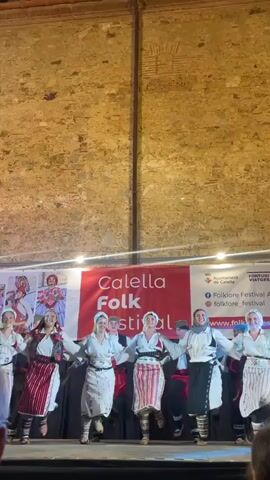 A group of approximately ten performers are on a stage in front of a brick wall.  They are holding hands in a semi-circle and appear to be dancing.  The performers are wearing traditional folk costumes.  Some of the women are wearing white blouses with embroidered vests and long, striped skirts.  Some of the men are wearing white shirts, vests, and darker pants or kilts.  Behind the performers is a large banner that reads "Calella Folk Festival".  To the right of the banner are smaller logos, including "Ajuntament de Calella" and "FORTUNI VIATGES".  The stage is lit by overhead lights, and some spotlights are casting red and white light onto the banner.  The overall environment suggests an outdoor evening performance at a folk festival.