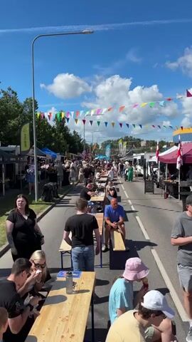 This is a vertical, eye-level shot of an outdoor street festival. The scene is bustling with people of all ages enjoying a sunny day. The street is lined with numerous market stalls and food trucks on both sides, adorned with colorful bunting strung across the road. In the foreground, several groups of people are seated at long wooden picnic tables, engaged in conversation and eating.

The subjects are primarily people, dressed in casual summer attire. Objects include market tents, food trucks, picnic tables, benches, and a street lamp. The setting is a street closure for a festival, likely a food or craft market, given the presence of stalls and food vendors.

The activity is a public gathering, where people are socializing, eating, and browsing the market. The time of day appears to be midday or early afternoon, indicated by the bright sunlight casting distinct shadows. The weather is clear and sunny, with a few scattered white clouds in a vibrant blue sky. The presence of bunting and the general atmosphere suggest a festive and joyful event.

Visible text includes the word "JÄRVE TALU" on a yellow sign on the left, which appears to be a vendor name. There are other signs on the market stalls and food trucks, but the text is too small or obscured to be legible. The overall emotion conveyed is one of community enjoyment and a lively, pleasant atmosphere. The location is confirmed as Tartu, Estonia.