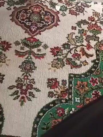 The video shows a close-up view of a rug with an intricate floral pattern. The rug has a cream-colored background with various floral motifs in shades of red, green, brown, and dark red. The pattern includes large central medallions and smaller floral clusters arranged across the surface. The texture of the rug appears to be thick and possibly hand-knotted. The camera pans slightly, revealing the edge of the rug with a green border. In the upper right portion of the frame, a small white object, possibly a piece of paper, is visible on the rug. The lighting suggests an indoor environment.