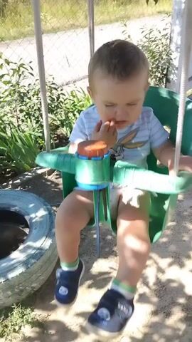 A toddler sits in a green swing. The child is wearing a light-colored t-shirt with a Bugs Bunny graphic and shorts. The child brings their hand to their mouth. On the ground below the swing are a tire, a small metal scoop, and an orange object. The swing is in a backyard with a chain-link fence and green foliage visible in the background. Sunlight casts dappled shadows on the ground.