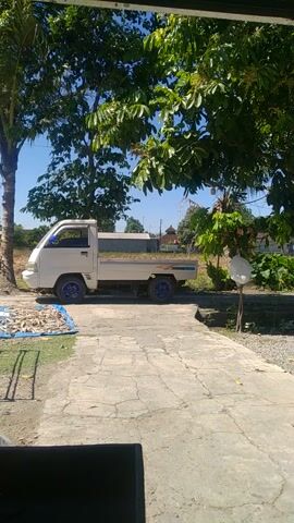 A white pickup truck with blue rims is parked on a concrete surface. Sunlight shines on the truck and surrounding foliage. The truck is seen from a side angle, and the background features trees, vegetation, and a clear blue sky. There are no people visible in the video.