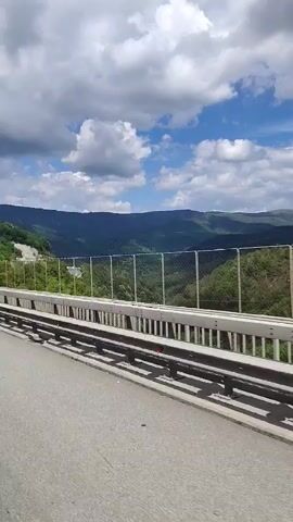 The video captures a scenic drive through a mountainous region on a clear, cloudy day. The camera is positioned inside a vehicle, looking out towards the landscape. The road surface is asphalt, with a metal guardrail running along the side. Beyond the guardrail, a chain-link fence separates the road from a densely wooded hillside. The trees are lush and green, indicating a time of year with vibrant foliage, likely spring or summer. The sky is a bright blue, dotted with fluffy white clouds that cast shadows on the rolling hills in the distance. The mountains appear layered, with shades of green and blue suggesting depth and distance.

A prominent blue sign is visible by the roadside, featuring text in both Cyrillic and Latin alphabets. The Cyrillic text reads "Община Ботевград" and the Latin text translates to "Botevgrad Municipality". This indicates the vehicle is passing into or through the administrative area of Botevgrad. The overall impression is one of a peaceful and picturesque journey through a rural, natural environment. The time of day appears to be late morning or early afternoon, given the brightness of the sky and the shadows. There are no people visible in the frame, and the focus is solely on the natural scenery and the road.