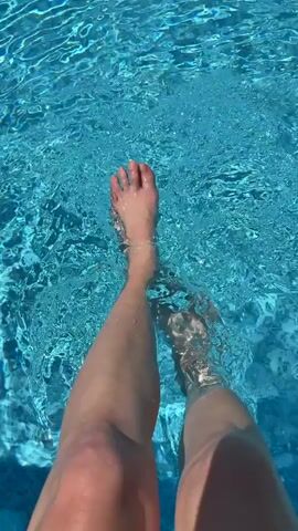 The video captures a first-person perspective of a person sitting by a pool, with their legs and feet submerged in the clear, blue water. The person gently moves their feet, creating ripples and small splashes on the water's surface. The sunlight reflects brightly off the water, enhancing its vibrant blue hue.