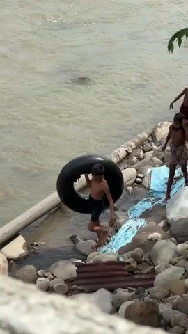 A young boy, wearing dark blue shorts, walks towards a muddy river carrying a large black inner tube over his right shoulder. He is on a rocky shore next to a concrete pipe. Behind him, another child, possibly a girl wearing a patterned skirt, walks towards the water. The river is wide and appears shallow with visible rocks and debris along the bank. The lighting suggests it is daytime.