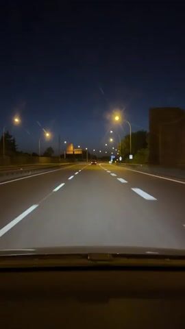 This video is a first-person perspective from inside a car driving on a highway at night. Streetlights illuminate the road, creating bright streaks of light. The sky is a deep blue, transitioning to black. Several cars are visible ahead, their taillights and headlights casting glows. Road markings, including white lines, are clearly visible. Trees line the side of the highway, and there are occasional signs and road infrastructure. The camera is positioned low, suggesting it's mounted on the dashboard.