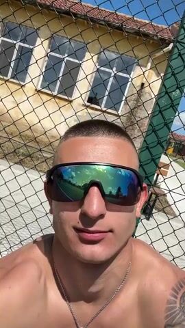 The video starts with a close-up of a shirtless man with a buzzcut, sunglasses, and a silver chain with a cross pendant standing in front of a chain-link fence on a sunny day. He is pointing his index finger upward. The camera then pans over a green sports field with white lines, revealing a soccer goal, a basketball hoop, and a husky dog. The camera focuses on a black harness with a chain leash and a blue rope toy on the grass. Finally, the camera returns to the man, who is still standing by the fence. The reflections in his sunglasses reveal the sports field. A building is visible behind the fence.