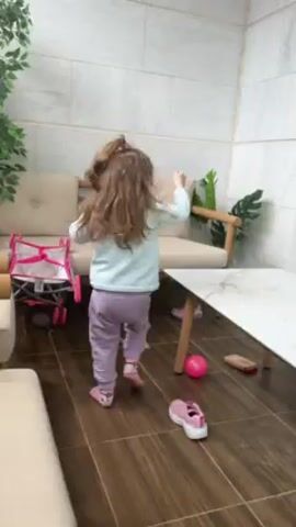 A young girl dances on a tiled balcony. She has brown hair pulled into a high ponytail and is wearing a light blue long-sleeved shirt, lavender pants, and pink shoes. She faces a sliding glass door, dances toward it, and turns to dance along the balcony to the left. The balcony has a white table with a marble-like top, a couch, and several plants. A pink ball and a single shoe are on the floor near the table. A pink toy stroller is next to the couch on the left. The walls are tiled and the sliding glass door has light curtains.