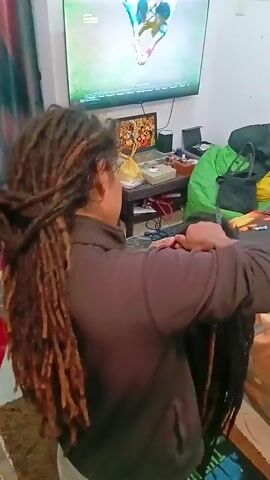 The video captures an indoor scene, likely a living room or studio, in Chopdem, India. Two individuals are present, with one actively styling the hair of the other. The stylist, seen from behind, wears a dark long-sleeved top and has numerous long dreadlocks extending down their back. They are focused on braiding or re-twisting the hair of the second person, whose back is towards the camera.

In the background, a large flat-screen television is mounted on the wall, displaying what appears to be a live sports broadcast, possibly cricket, featuring several male figures on a green field. Small numbers like "1.74" and "9.99" are visible on the screen, typical of a sports match overlay. Below the TV, a small table holds a framed picture, a yellow plastic bag, and other items. To the right, another cluttered table features various objects, including potential hair products or accessories. A green beanbag chair, a black shoulder bag, a red-covered stool, and other bags are also visible around the room. The floor is covered with patterned tiles. The scene conveys a casual and lived-in atmosphere, with the activity taking place under indoor lighting.