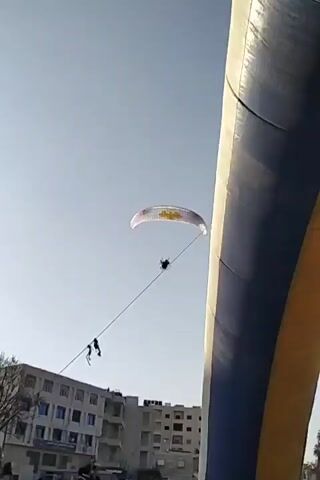 A person flies a powered paraglider in a blue sky with a Syrian flag visible. The camera view is partially obstructed by a large blue and gold archway. The paraglider has a white canopy with yellow markings. A building is visible in the background.
