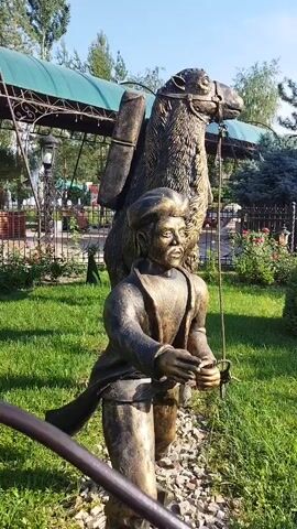 A metal sculpture of a man leading a camel is displayed outdoors. The sculpture depicts a man in traditional clothing holding the camel's reins. The camel is loaded with cargo on its back. The sculpture is situated in a grassy area with some plants and flowers. In the background, there is a green awning, a fence, and buildings, suggesting a park or an outdoor recreational space. The lighting indicates it is daytime, likely sunny.