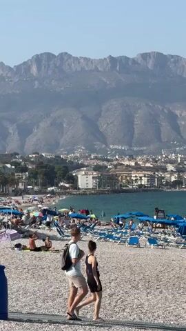 This media file captures a lively scene at a pebble beach in Pla Parcial Platja, Spain. The foreground is dominated by a couple walking along the shore, the man with a backpack and the woman in a black dress. Behind them, the beach is dotted with numerous blue sun loungers and umbrellas, indicating a popular spot for sunbathers. Many people are visible lounging, walking, or engaging in activities on the beach.

The middle ground features the turquoise waters of the sea, with several boats anchored offshore. Beyond the beach and the sea, a bustling town is nestled against the backdrop of imposing, rugged mountains. The town displays a mix of residential buildings and some larger structures, one of which appears to be a church with a distinct dome. The overall impression is one of a vibrant coastal resort town.

The time of day appears to be late morning or early afternoon, judging by the bright sunlight and the long shadows cast on the beach, suggesting pleasant weather. The clear blue sky with a few wispy clouds adds to the summery atmosphere. The emotions conveyed are relaxed and happy, typical of a beach holiday. There is no visible text in the media file.