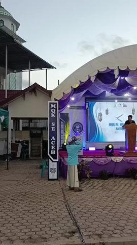 A staged event is set up outdoors. There is a large tent with purple and white drapery. In the center, a digital screen displays graphics and text. To the right of the screen, a person stands at a wooden podium wearing dark clothing and a head covering. To the left of the stage, a tall, vertical sign displays text in white and blue. In front of the sign, a person wearing a light blue shirt and white pants stands facing the stage. Further to the right and in front of the stage, a child in a yellow shirt runs across the paved ground. Several spotlights are positioned on the stage. In the background, there is a building with a mosque-like structure on the left side. The sky is overcast.