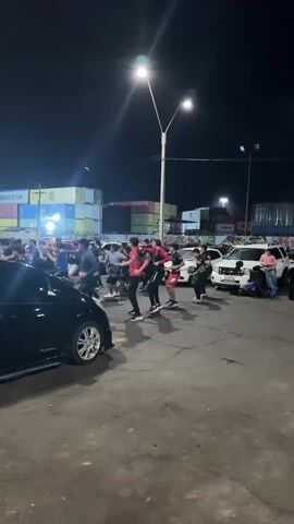 The video shows a group of young men dancing outdoors at night. They are in a paved area with shipping containers in the background and cars on the side. The men are wearing casual clothing, some in matching red shirts with black accents. They are performing synchronized dance moves, including jumps, kicks, and arm movements. Some of the dancers are holding circular objects, possibly drums or tambourines, which they are playing as part of the performance. The lighting is provided by streetlights, casting shadows on the ground. The overall atmosphere appears energetic and festive.