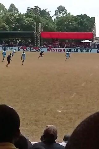 The video shows a soccer game. Players in black and light blue uniforms are playing on a dry grass field. A banner that reads "WESTERN BULLS RUGBY" is along the edge of the field. The scene is captured from the stands where spectators are seated. There are tents in the background.