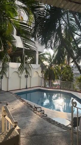 The camera looks out on a swimming pool in an outdoor area, which is partially shaded by palm trees. The pool is surrounded by a stone patio. A white two-story building is on the left. It has arched decorative features on the ground floor and appears unfinished on the second floor. A white metal fence runs along the back of the pool area. Palm trees and other plants are visible beyond the fence. The sun is reflected on the surface of the water. A few chairs are visible around the pool. There is a white railing in the foreground, suggesting the camera is positioned on a balcony or elevated area.