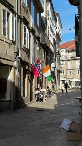 A narrow, sun-drenched cobblestone street in Santiago de Compostela, Spain, is lined with tall, old buildings featuring stone facades and numerous windows. On the left, a shop displays a deep red garment in its window. Two flags, Norway and India, hang prominently from a building, adding a splash of color to the scene.

In the foreground, several people are seated at tables outside a cafe, enjoying the sunshine. They appear relaxed, with one person reading and others engaged in conversation. Further down the street, more people are seated at outdoor tables, suggesting a lively atmosphere. A person is walking a dog, adding a touch of everyday life to the scene.

The sunlight casts strong shadows on the cobblestones, indicating it is a bright, sunny day. The clear blue sky overhead suggests pleasant weather. The architecture of the buildings, with their balconies and traditional stonework, points to a historic European city. The presence of outdoor seating and people enjoying themselves creates a feeling of leisure and community.

A sign on the right side of the street is partially visible, with some text in white on a dark green background. Further down, a sign with yellow lettering on a dark background is also visible. In the lower right corner, a few cardboard boxes are stacked, suggesting deliveries or preparation for operations. The overall impression is one of a charming, vibrant European street on a beautiful day.