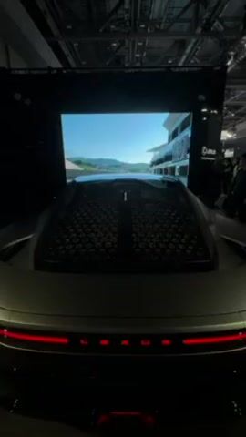 The video shows the back of a silver Lexus car, viewed from a slight angle. The rear window has a unique, patterned design of closely-packed circular shapes. A large screen behind the car displays a racing simulator game, showing a track with buildings and scenery passing by. The screen also features the word "Lexus" in the upper-right corner and "Buble" near the bottom-right. Red LED taillights are visible on the car, with the letters "LEXUS" illuminated between them. People can be seen standing around the car, particularly on the right side, suggesting an event or exhibition setting. The environment appears to be a large, indoor venue with a high ceiling and visible support beams.