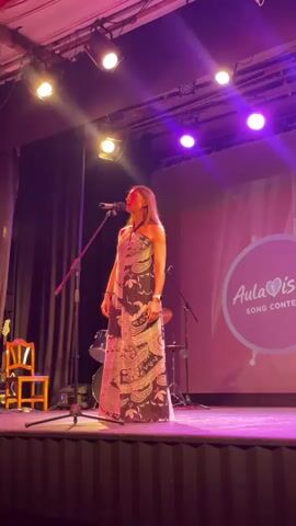 A woman stands on a stage singing into a microphone. She is wearing a long, patterned dress. There are stage lights above her and a backdrop with text that says "AulaDis Song Contest". There are also musical instruments visible on the stage.