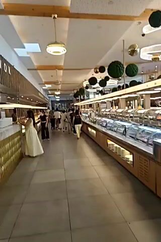The video begins with a close-up of a light gray tiled floor. The camera pans down a hallway with the same tile. A pair of light brown slip-on shoes is visible in the lower center of the frame. The camera continues to move forward, showing the shoes and the tiled floor. At the end of the video, a finger briefly enters the frame in the foreground. The overall environment appears to be indoors, possibly a commercial space like a store or a mall, given the polished tile and the appearance of store fixtures in the background at the beginning of the video.
