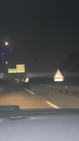 The video, taken from inside a vehicle at night, shows a highway scene. There are several signs visible above the road, indicating directions to CHARLEVILLE-M, AEROGARE, ST-REMI-CHAMPAGNE, ST-BRICE-COUCY, PARC D'EXPO, and Z.A. DU CHATELET-MAURIZIENNE. In the distance, there are several figures in yellow vests, presumably workers or emergency personnel, illuminated by vehicle lights. A yellow circular sign with a black border is also visible on the side of the road. The overall environment is dark, with artificial lighting from street lamps and vehicle headlights.