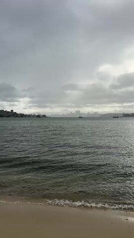 A vertical video shows a view of a body of water from a sandy beach. The sky is overcast and gray, with visible clouds. Gentle waves are lapping at the shore. In the distance, across the water, there is a hazy shoreline with buildings and structures. Two sailboats are visible in the middle ground of the water. The overall atmosphere is subdued and appears to be a cloudy day.
