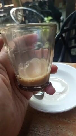 Here's a detailed description of the video:

A hand holds a nearly empty, clear glass that contains a small amount of light brown liquid, likely coffee or tea, at the bottom. The drink has some foamy residue on the side. The glass sits on a small white saucer, with a teaspoon to the side. There appears to be a small puddle of liquid on the saucer as well.

The background is slightly blurred, but a wooden table is visible beneath the saucer. Beyond that, there's a black metal chair and a motorcycle partially visible in the distance. The overall lighting suggests an outdoor setting, likely during daylight.
