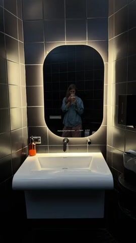 A person is filming a video in a bathroom. The bathroom has black tiles on the walls and a sink with a mirror above it. The mirror is illuminated with LED lights around its edge. The person is taking a selfie in the mirror with their phone. The video pans up to show a ceiling with a graphic of outer space, featuring stars and a planet. The lighting in the bathroom is dim, with some bright spots from the mirror's illumination. The person is wearing a light blue shirt and a pink skirt.