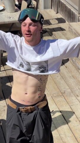 A man in a cropped t-shirt, black pants, and a brown belt stands on a wooden deck. He wears goggles on his head and has several tattoos on his torso. The camera zooms in on his abdomen, focusing on a tattoo that reads "love always." The man then walks away, and the camera follows him as he walks onto a crowded outdoor patio. People sit at tables and walk around, many dressed in ski gear. The video ends with a view of a DJ mixing board, suggesting music is being played.