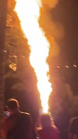 A crowd of people stands on a beach at night, watching a performer shoot a large column of fire into the air from a device held above their head. The flames are bright orange and yellow, and smoke billows around them. The people in the crowd are holding up their phones to record the performance. String lights are visible in the background, and palm trees are silhouetted against the night sky. The performer is wearing a red dress, and the people in the crowd are wearing casual clothing. The scene appears to be a tourist destination.