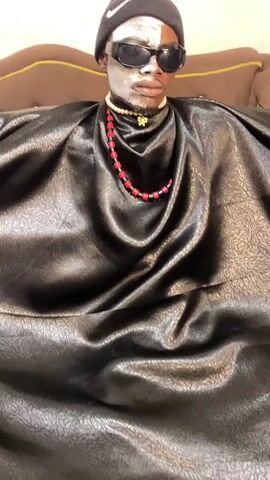 A person wearing a black beanie and black sunglasses with white face paint on their face, and a beaded necklace, sits on a couch. They are covered by a large piece of black material.