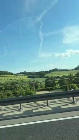 This is a first-person perspective video taken from inside a car driving on a highway on a clear, sunny day. The video pans across the landscape, showing green rolling hills, trees, and a blue sky with some wispy clouds. The car passes by a highway guardrail and a blue sign with German text, indicating an exit. A side mirror shows the reflection of the car and the road behind. The dashboard of the car is visible in the foreground, with some small cartoon figures attached to it.