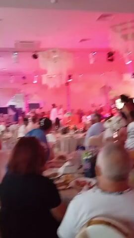 The video shows an indoor event, possibly a wedding reception or a formal party, with guests seated at round tables decorated with white tablecloths, flowers, and chairs adorned with sashes. The room is illuminated with pink and purple lighting, and decorative elements hang from the ceiling. In the background, a stage area is visible with what appears to be a band or performers. People are seen walking around, interacting, and some are dancing. The camera is positioned from the perspective of a seated guest, looking towards the stage and the general activity of the event.