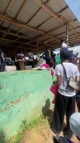 The video shows an outdoor gathering of people, many of whom are wearing white clothing. The scene appears to be a community event or a formal gathering, possibly a school or organization event. There are structures with roofs providing shade, suggesting a sunny day. People are seen standing, walking, and interacting. Some individuals are holding bags or papers. In the background, a large group of people in white attire are lined up or gathered in a field. There are trees and an open sky with clouds visible. The ground is a mix of dirt and some grassy areas. Some areas appear to have makeshift stalls or gathering points under the shaded structures. The overall impression is of a busy event with many participants.