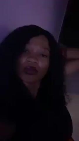 A woman with dark hair and lipstick is in the video. She wears a black sleeveless top and an orange and black patterned skirt. The background is mostly a light purple wall. The lighting is dim.  The woman moves slightly, adjusting her hair at one point.