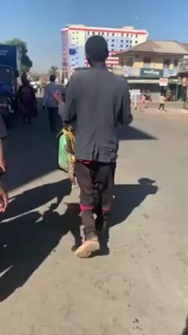A man wearing a black blazer, black pants with red stripes, and beige sandals is walking on a street while pushing a wheelbarrow laden with items covered by a green bag. The scene is set in a busy urban environment during daylight. Other pedestrians are walking on the sidewalk and street alongside vehicles, including a large truck with "Klin CLEANING POWER" written on its side. Buildings are visible in the background, one of which is tall and painted in blue, red, and white. The sun casts shadows on the street, suggesting a bright and sunny day.