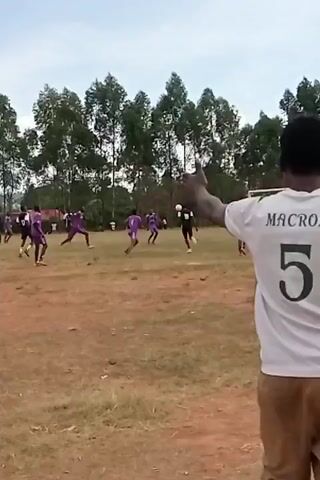 A soccer game is being played on a grassy field. Players in purple jerseys and players in black jerseys are competing. The soccer goal is visible on the right side of the frame. A line of tall trees is in the background. A person in a white shirt is standing near the camera on the right. The person is partially visible. Their arm is raised. The weather appears to be cloudy and overcast.