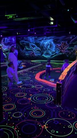 The video shows a family playing mini-golf in a dark room illuminated with blacklights. The room has glow-in-the-dark carpet with circle and star designs, and blacklight-painted murals of sea creatures on the walls. People, including adults and young children, are scattered around different mini-golf holes, moving around the course. A woman in white stands near a child at one hole, while other children are near another hole. Some mini-golf holes have obstacles like a shark or cannon.