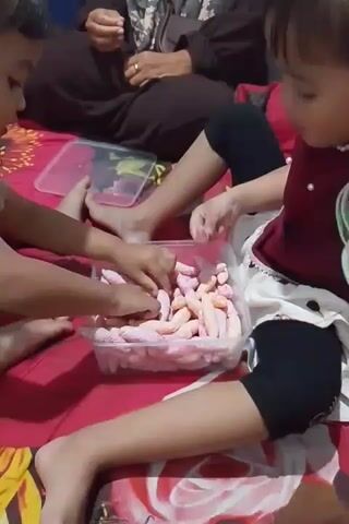A group of children and an adult are gathered on a bed with a red and white floral print. In the center of the frame, a clear plastic container is filled with numerous small, elongated pink and white snacks. Two children are actively engaged with the snacks. One child on the left is reaching into the container with both hands, picking up several snacks at once. To the right, another child is holding a snack up to their mouth, seemingly about to eat it. Their hands are near the container. A third child, positioned between the two, is also interacting with the snacks, with their hands near the container and their head bent down. The adult in the background, wearing a dark brown garment, is partially visible, with their hands resting in their lap. The lighting is dim, suggesting an indoor setting.