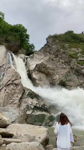 A video shows a powerful waterfall cascading down a rocky cliff face. Two young women are visible in the foreground, standing on large rocks near the base of the waterfall, facing away from the camera and towards the rushing water. One woman is wearing a white shirt and a long white skirt, and the other is wearing a light-colored garment. The surrounding environment is rugged and natural, with lush green trees and vegetation growing on the slopes above the waterfall and along the cliff edges. The water in the river below the waterfall is turbulent and frothy, creating a mist that hangs in the air. Several large rocks are scattered in and around the riverbed. In the distance, on a higher vantage point of the cliff, a small group of people can be seen observing the waterfall. The sky is overcast with a cloudy sky.