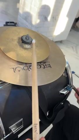 A close-up POV shot captures a person playing a drum set. The focus is on a drumstick about to strike a cymbal. The drumstick is made of light-colored wood with the brand name "IRIN" visible. The cymbal is golden-colored with text printed on it, including "Istanbul". Another drumstick is visible in the background, being held by another person's hand. The drum set itself is black. The background is blurred but appears to be an indoor setting with bright lighting and a hint of a brick wall and a person's shadow. The audio consists of drumming sounds, specifically cymbal strikes, accompanied by singing.