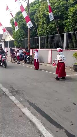 A street scene with a paved road and a sidewalk. Several people are present. On the road, a motorcycle with two riders is moving from left to right. Two young females in red skirts and white headscarves, likely students, are walking from left to right on the sidewalk, facing slightly towards the camera. Another young female in a similar outfit, carrying a pink backpack, is standing near a parked motorcycle on the left side of the road. In the background, several people are gathered near parked motorcycles and a bicycle. The environment is decorated with Indonesian flags, red and white pennants strung from a pole and attached to a building. Trees and a residential building are visible in the background. The overall atmosphere appears to be a procession or a community event.