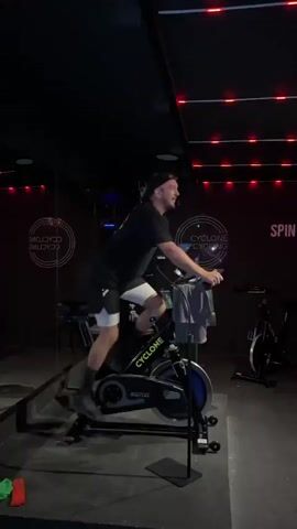 A man is riding a stationary exercise bike in a dimly lit room. He is wearing a black t-shirt, white shorts, black socks, and a baseball cap. He glances towards the camera and smiles. The exercise bike is black and has the word "CYCLONE" printed on it. A towel is hanging from the handlebars. The room has dark walls and a mirrored wall with the words "CYCLONE CYCLING" in a circular logo. There are other stationary bikes visible in the background. Red lights are visible on the ceiling. The lighting in the room alternates from very dark to bright. He seems to be leading an exercise class.