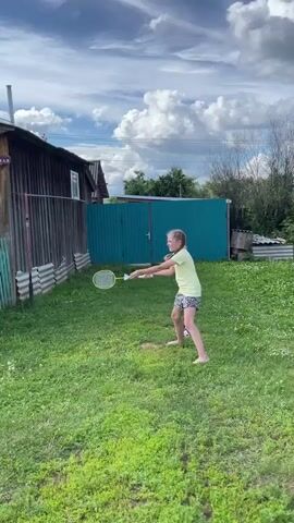 A young girl with blonde hair is playing badminton in a grassy yard. She is wearing a yellow t-shirt and patterned shorts. The girl is in motion, swinging a badminton racket. She is barefoot. In the background, there is a wooden building with a fence. The sky is cloudy.