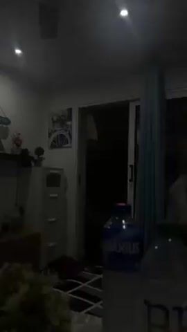 The video shows an indoor scene in a dimly lit room. The lighting appears to be provided by overhead lights. Two water bottles, one with a blue label ("Aquarius") and the other with a green cap, are prominently placed in the foreground. A dark-haired person, wearing a black long-sleeved shirt, briefly comes into view, manipulating an object, possibly a hair tie. In the background, there is a sliding glass door covered by a light blue curtain, along with a white cabinet holding various decorative items. A painting featuring a bicycle is visible on the wall. The general atmosphere suggests an informal, domestic setting.