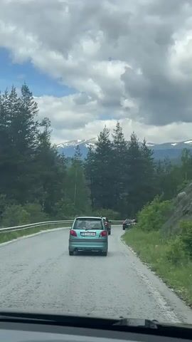 A blue car is driving on a road. There is a motorcycle behind the car. There are trees on both sides of the road. There are mountains in the background. The sky is cloudy.