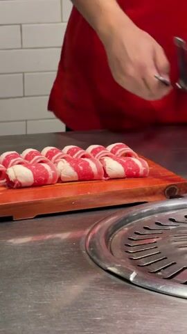 A person wearing a red shirt is preparing thinly sliced meat rolls on a wooden cutting board. The meat appears to be beef, with alternating layers of red meat and white fat. The person is using silver tongs in their left hand to hold a piece of meat while using scissors in their right hand to cut it. Several other meat rolls are visible on the board, arranged in rows. The cutting board is placed on a stainless steel surface, which has a round grill grate in the foreground. The background shows a white tiled wall. The lighting is bright and seems to be from overhead.